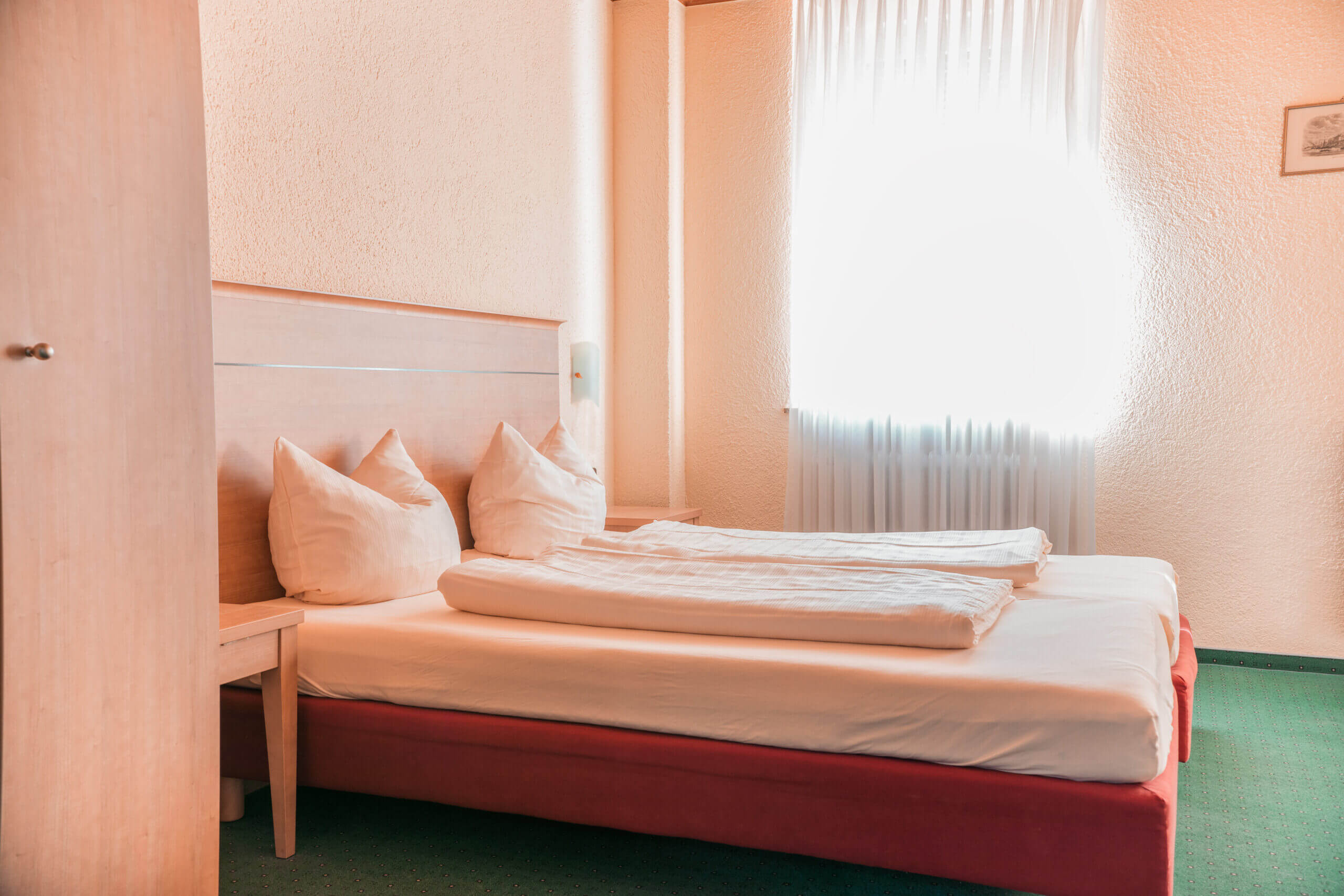 Ein gemütliches Hotelzimmer im Hotel St. Paulushof mit einem Doppelbett, hellem Holzdekor, weißer Bettwäsche und einem hellen Fensterbereich.